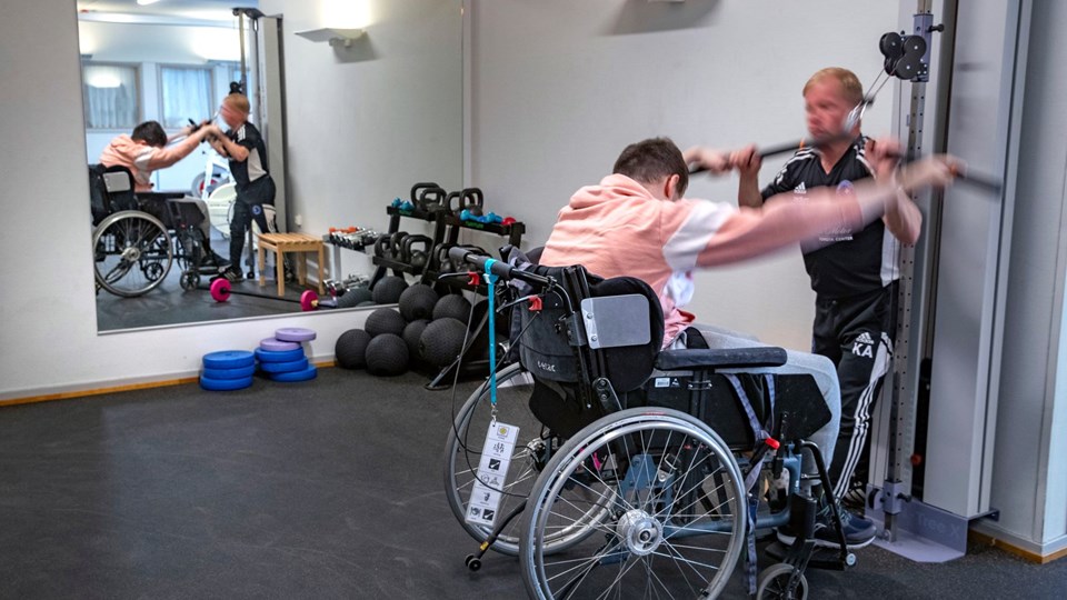 Elev i rullstol tränar tillsammans med vuxen i ett rum med träningsredskap.