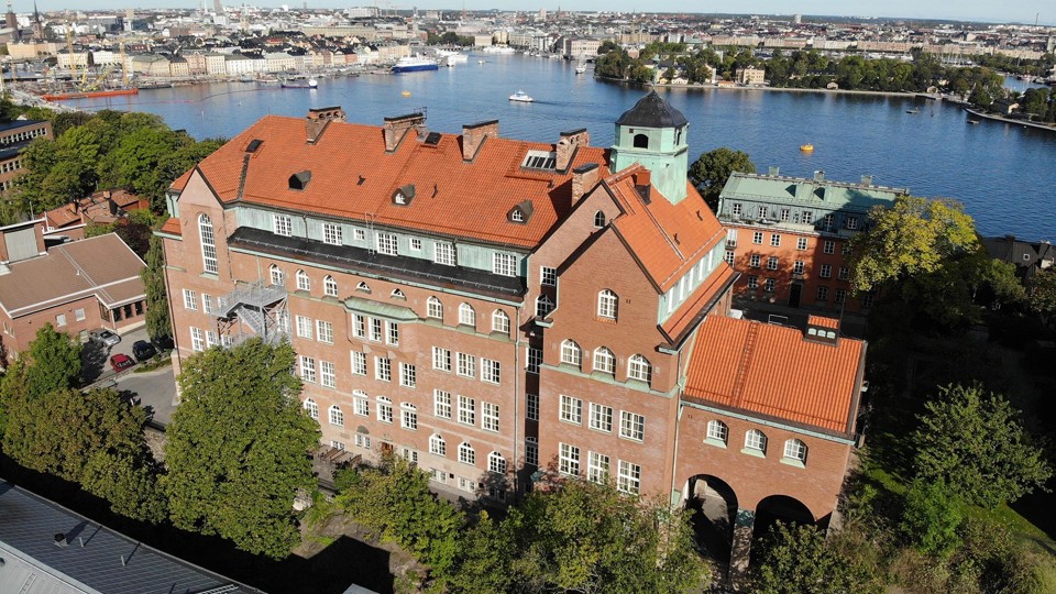 P A Fogelströms gymnasium ses snett uppifrån. Bakom skolar syns delar av Stockholm och Stockholms vatten, bland annat Gamla stan, Strandvägen, Djurgårdsfärjan och andra båtar och fartyg.